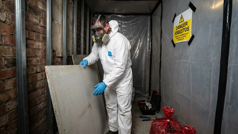 Worker in protective equipment removing asbestos insulating board from a UK building